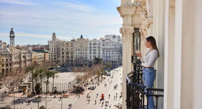 Mulher em sacada de prédio observando vista panorâmica de centro urbano com arranha-céus, prédios históricos e movimentação de pedestres na praça abaixo, no hotel Venecia Plaza Centro. Representa onde ficar em Valência.