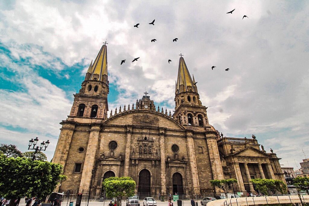 Catedral de Guadalajara, um grande edifício religioso de arquitetura colonial espanhola com torres gêmeas douradas em um céu nublado, com bandos de pássaros voando acima.