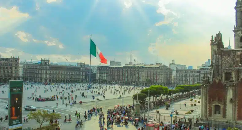 Zócalo com multidão de pessoas, bandeiras nacionais tremulando, prédios históricos ao redor e céu azul com nuvens, para representar o que fazer na Cidade do México.