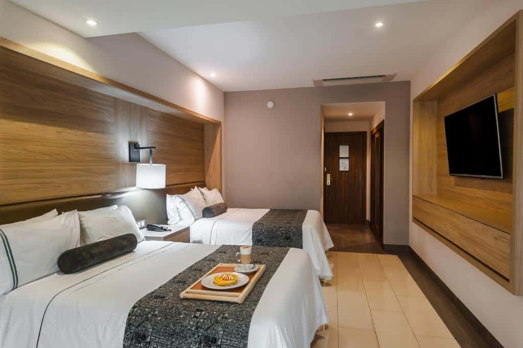 Quarto moderno no Hotel Real Maestranza com duas camas de casal, televisão de tela plana na parede, e mesa de centro com bandeja de café da manhã, para representar onde ficar em Guadalajara.