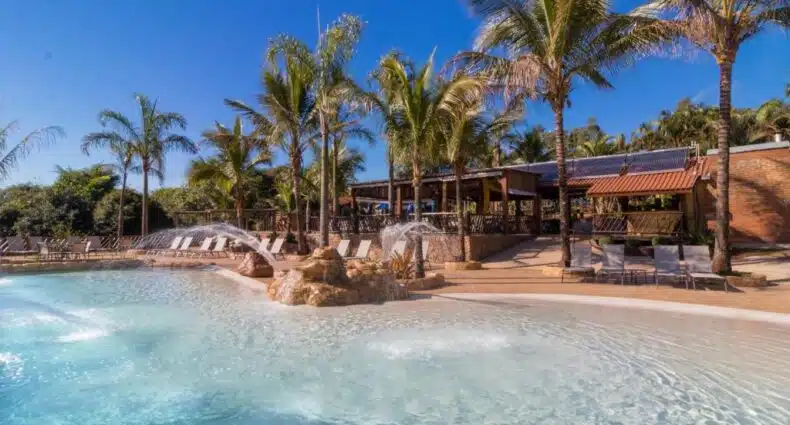 Piscina do Recanto Alvorada Eco Resort com palmeiras ao fundo, com espreguiçadeiras ao redor da borda da piscina. Imagem para ilustrar post sobre hotéis fazenda e resorts em Brotas.