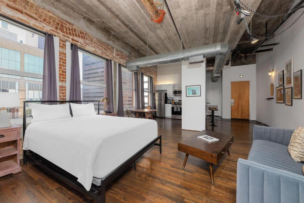 Quarto de loft industrial moderno com paredes de tijolo aparente, teto com tubulações expostas e grandes janelas com vista para a cidade. A decoração inclui uma cama de casal branca, uma mesa de centro de madeira vintage, um sofá cinza e diversos quadros nas paredes.