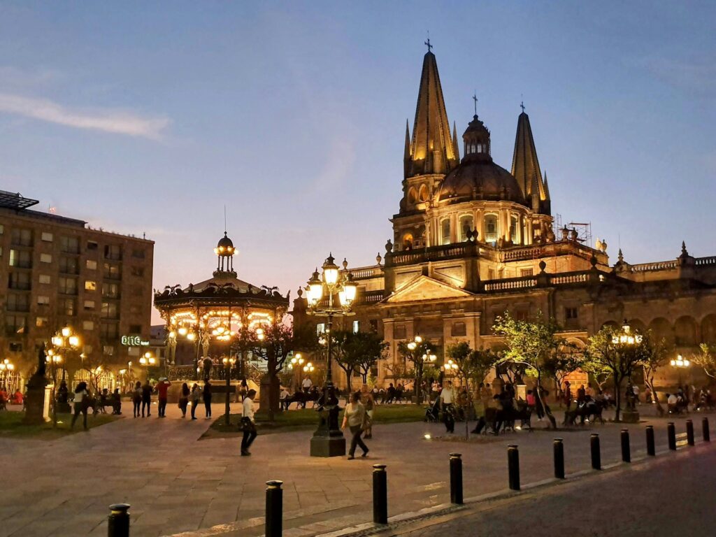Praça da Catedral Metropolitana de Guadalajara à noite, com cúpula e torres iluminadas brilhantemente contra o céu escuro, rodeada por pessoas caminhando e atividades na praça.