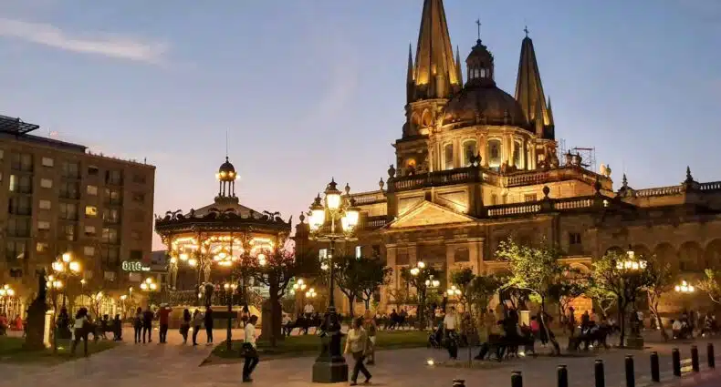 Praça da Catedral Metropolitana de Guadalajara à noite, com cúpula e torres iluminadas brilhantemente contra o céu escuro, rodeada por pessoas caminhando e atividades na praça, para representar onde ficar em Guadalajara.