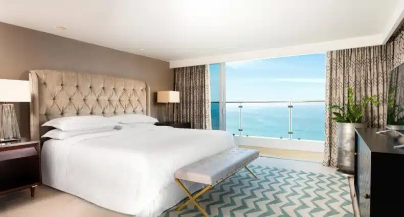 Quarto de Sheraton Grand Rio Hotel & Resort com vista para o mar, com cama de casal de estofado cinza e detalhes dourados, cortinas e tapete com padrão geométrico, e paisagem do oceano ao fundo. Imagem para ilustrar post sobre resorts all inclusive no Rio de Janeiro.