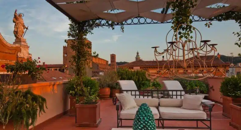 Varanda ensolarada com vista panorâmica da cidade, com estátua, telhados e torre de igreja ao fundo, decorada com lustre de ferro forjado, sofá e mesa de centro em área externa aconchegante, do Grand Hotel Majestic gia' Baglioni.