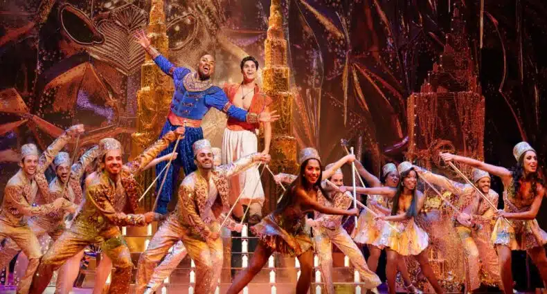 Elenco do musical Aladdin em palco com figurinos brilhantes e cenário oriental dourado