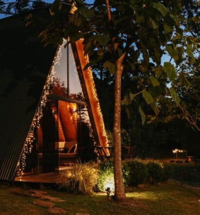 Casa de campo moderna com telhado em formato de triângulo iluminada à noite por luzes decorativas entre as árvores.