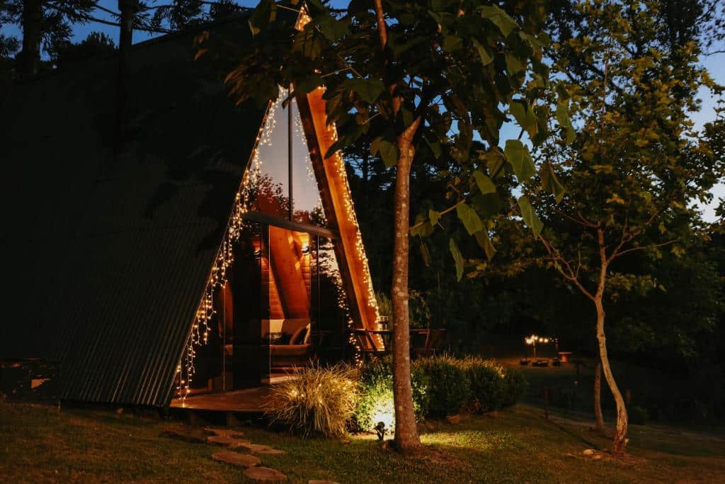 Casa de campo moderna com telhado em formato de triângulo iluminada à noite por luzes decorativas entre as árvores.
