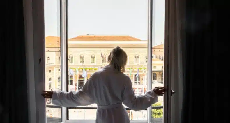 Mulher de roupão branco no Mercure Bologna Centro observando a vista da janela de um prédio com fachada histórica em centro de cidade. Representa onde ficar em Bolonha.