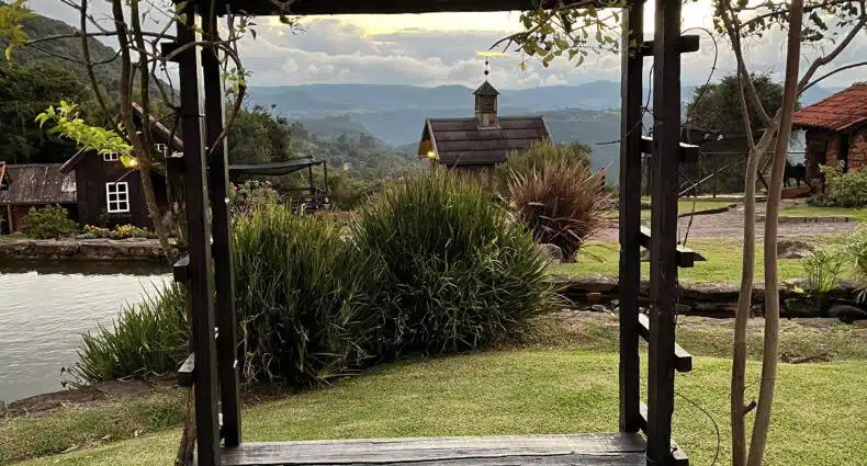 Pérgola de madeira escura com banco em jardim gramado com vista para montanhas e lago