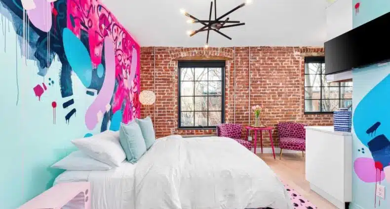 Quarto amplo da The Acoma House com parede decorada com grafite colorido, mobiliário moderno e iluminação pendente preta em estilo industrial, com vista para jardim através de grandes janelas. A imagem ilustra o post sobre onde ficar em Denver.