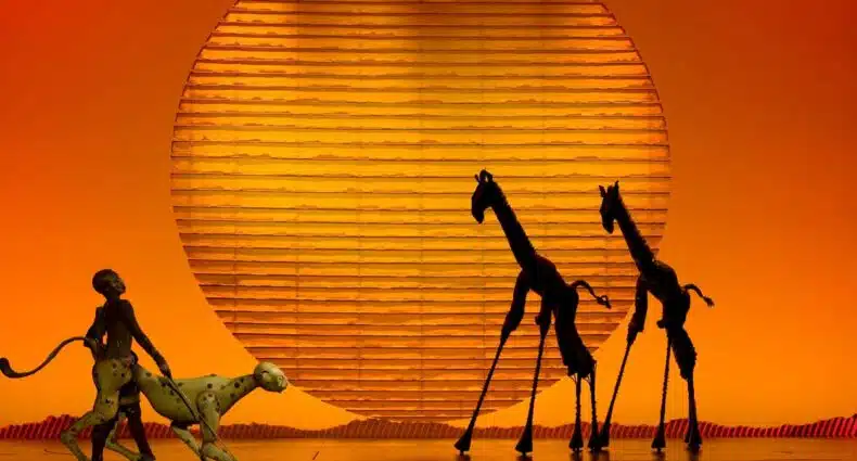 Cenário do musical O Rei Leão com silhuetas de Simba, Timão e Pumba sob sol laranja no palco