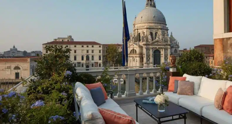 Praça em Veneza, Itália, com a famosa Basílica de Santa Maria da Saúde no fundo, vista de um terraço com móveis de estar brancos e laranja.