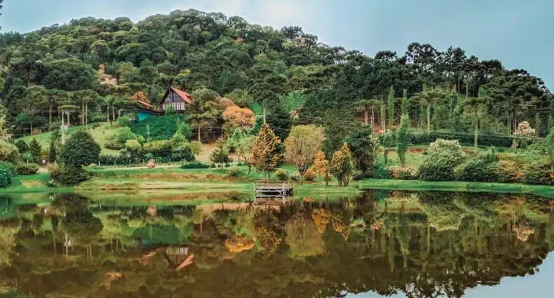 Casa de campo de madeira em Urubici refletida em lago verde rodeado por vegetação em um cenário de montanhas. A foto é capa do post de cabanas em Urubici.