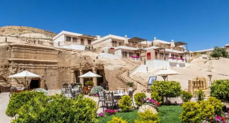 Edifício do Petra Guest House Hotel de estilo árabe com varandas, em cenário de jardim com vegetação exuberante e mesas de café ao ar livre. Representa o post onde ficar em Petra.