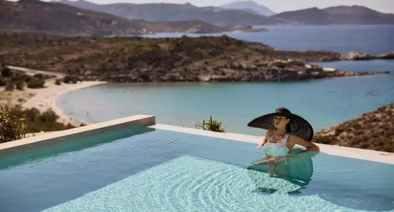 Mulher relaxando em piscina com vista para praia paradisíaca e mar azul turquesa no Hotel Milos Sea Resort. Representa onde ficar em Milos.