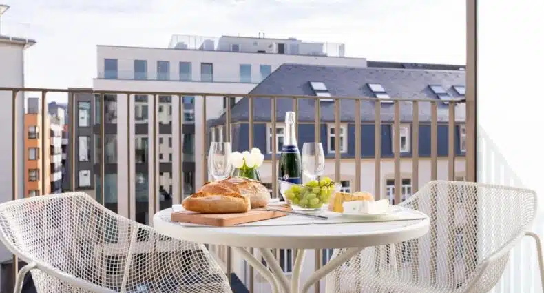 Varanda de apartamento com mesa redonda, pães, queijos, uvas e champanhe para café da manhã romântico no The Central City. Representa onde ficar em Luxemburgo.