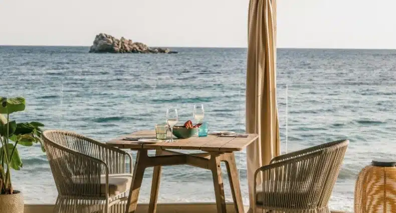 Mesa de jantar em restaurante à beira-mar com vista para ilha rochosa e mar azul em dia ensolarado