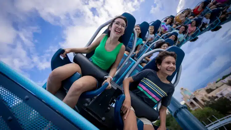 Pessoas sorrindo em montanha-russa com céu azul ao fundo, diversão em parque de diversões