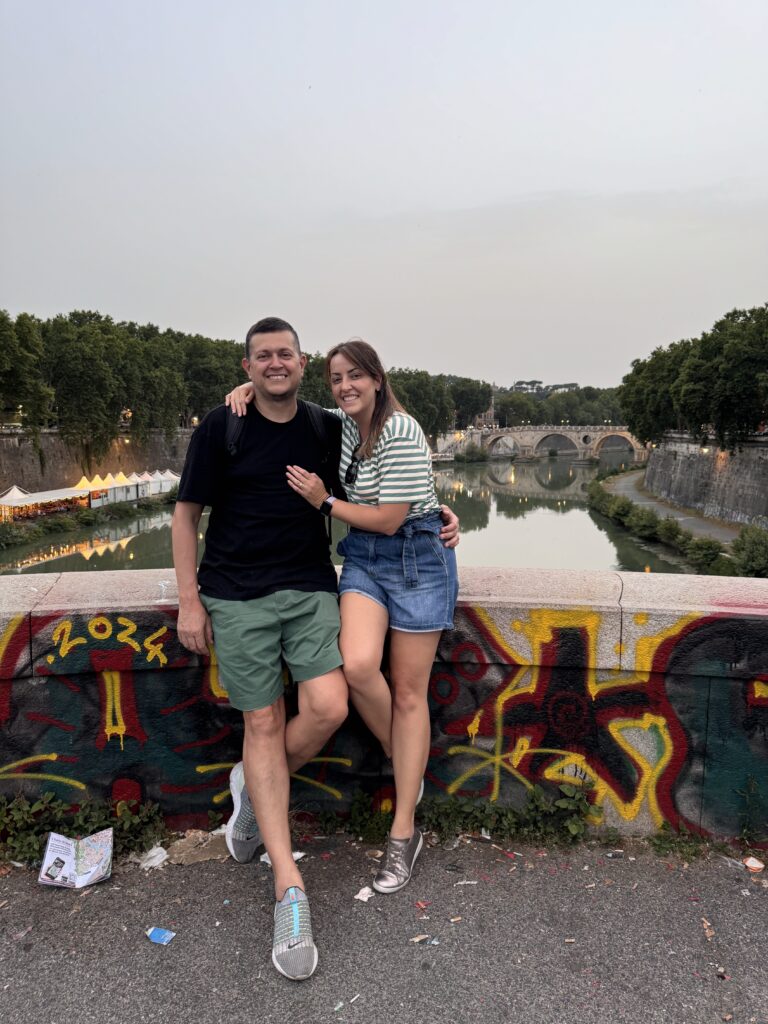 Flávio e Juliana sorrindo em frente ao rio Tibre e Ponte Sant'Angelo em Roma, Itália