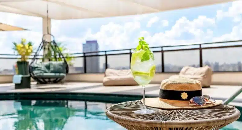 Coquetel refrescante com limão e hortelã ao lado de chapéu de palha e óculos de sol em piscina de hotel com vista para cidade do Grand Mercure SP Itaim Bibi. Representa hotéis em São Paulo.