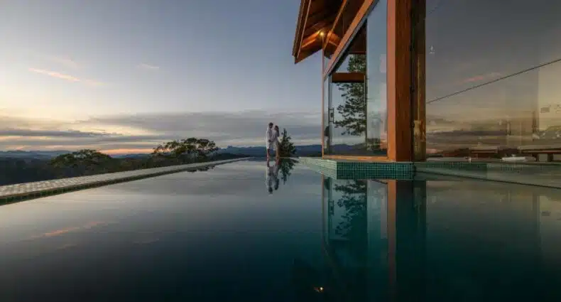 Casal admirando pôr do sol em piscina infinita com vista para montanhas na natureza brasileira do Quinta dos Pinhais. Representa pousadas românticas no interior de SP e sul de Minas.