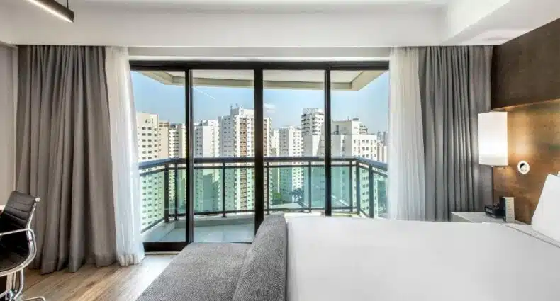 Quarto de hotel moderno com vista panorâmica da cidade de São Paulo, cama de casal e poltrona cinza