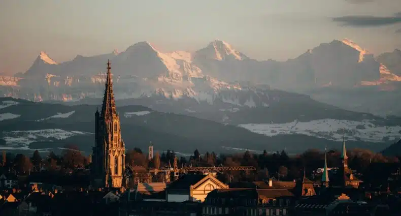 Paisagem de Berna com montanhas nevadas dos Alpes Suíços ao fundo e catedral com torre alta. A imagem ilustra o post sobre onde ficar em Berna.