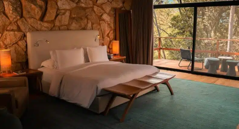 Quarto do Botanique Hotel Experience com cama de casal, decoração rústica e vista para área verde com varanda e cadeiras. Representa hotéis para fim de semana em São Paulo.