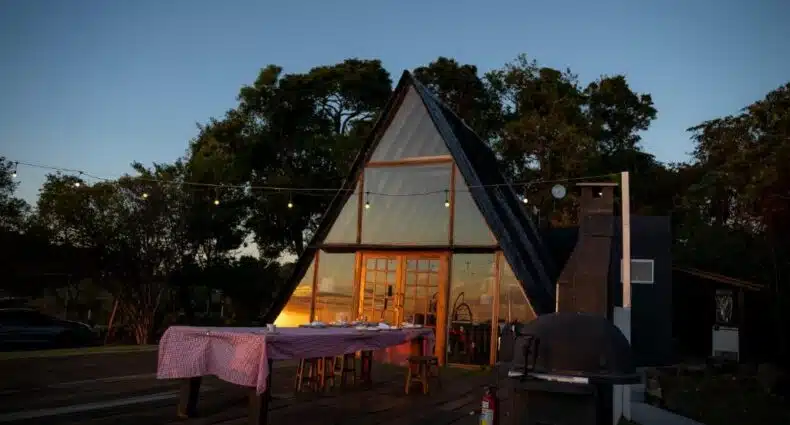 Casa triangular de madeira com deck e mesa posta para jantar ao ar livre com luzes da Cabana Pico do Gavião. Representa chalés em Minas Gerais.