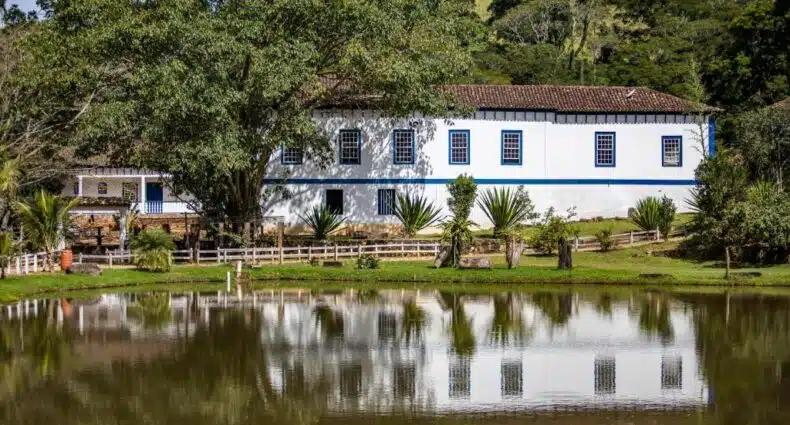 Fazenda histórica colonial branca e azul refletida em lago com vegetação exuberante no Hotel Fazenda Paciencia. Representa hotéis fazenda para o Réveillon em MG.