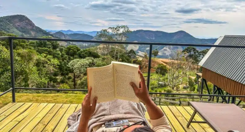 Mulher lendo livro em deck de madeira da Cabana Encanto do Sapucaí com vista para montanhas verdes, em São Bento do Sapucaí. Representa um dos chalés na montanha em SP