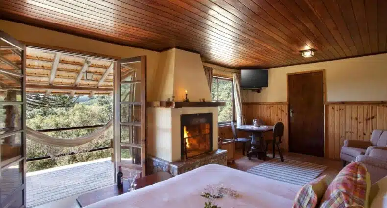 Quarto do Hotel Fazenda Itapuá com lareira, cama de casal e vista para área verde com rede. Representa hotéis fazenda baratos em MG.