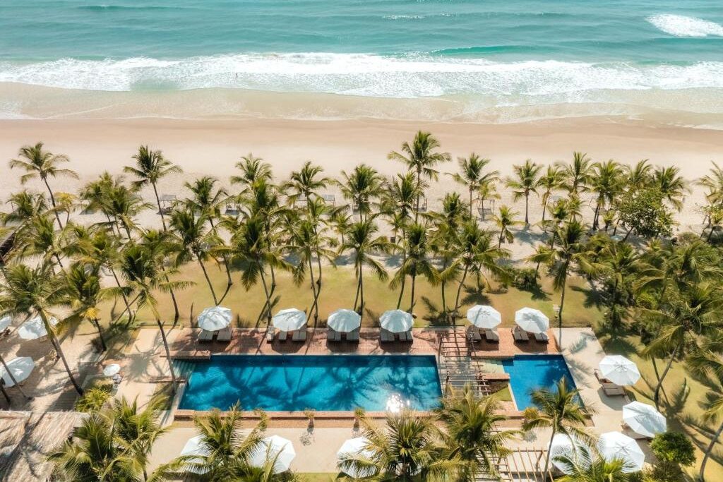 Piscina retangular azul no Txai Resort em frente a praia com coqueiros e guarda-sóis brancos.